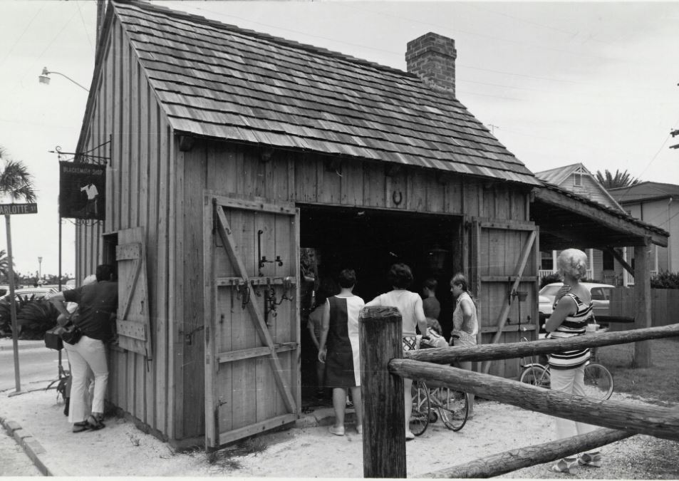 A black and white photograph of people watching a live demonstration at a wooden blacksmith shop.