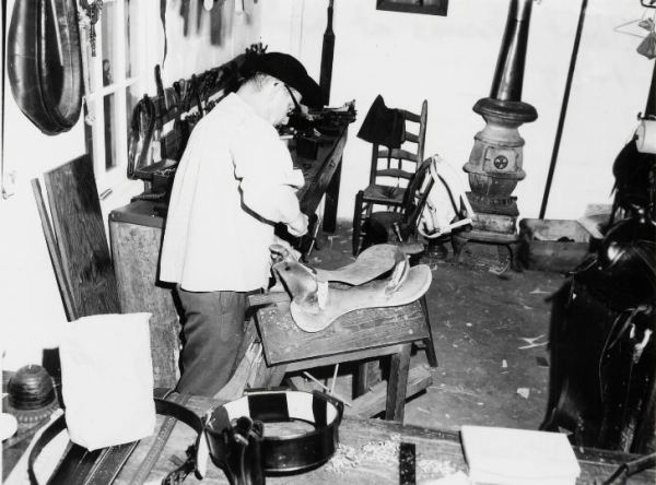 A black and white photograph of Albert Thomas dressed in a colonial costume working on a saddle in a leather shop.