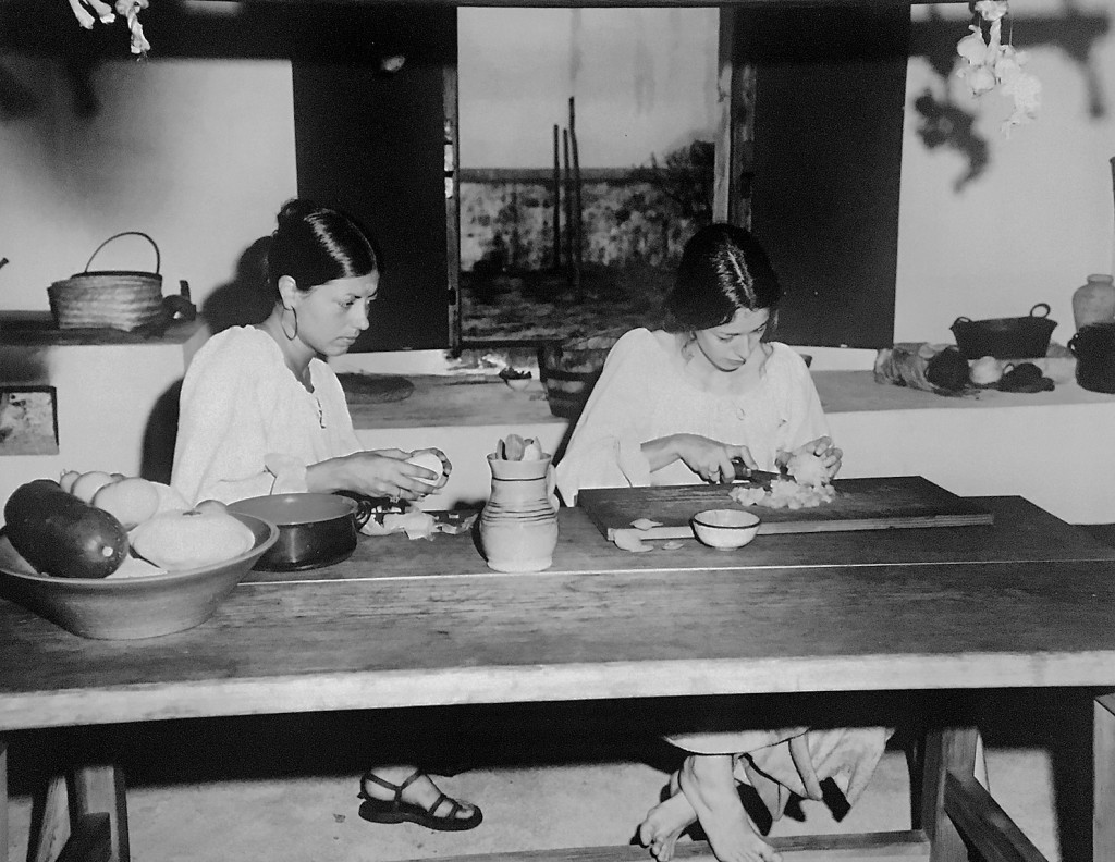 A black and white photograph of two women preparing food at a wooden table in a kitchen.