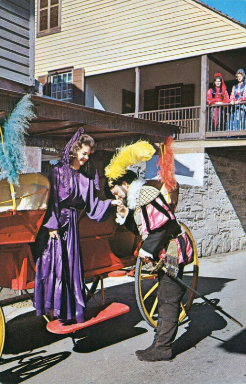A color photograph of a man dressed in Spanish colonial inspired costume kissing the hand of a woman in a Spanish colonial inspired costume exiting a carriage. Two women in Spanish colonial inspired costumes look on from a balcony in the background.