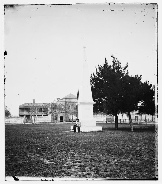 A black and white photograph of a person sitting on the base of an obelisk in a field with buildings in the background.